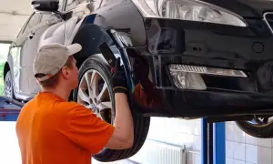 Mechanik w pomarańczowej koszulce sprawdza błotnik czarnego auta na podnośniku. Może to być powód, dlaczego mechanicy w dużych aglomeracjach rezygnują z napraw "od ręki".