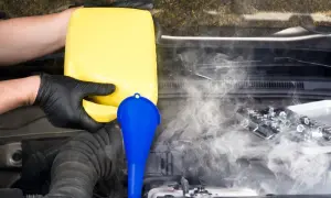 Ręce w czarnych rękawiczkach wlewają żółty płyn do silnika przez niebieski lejek. Para unosi się, sugerując problem, dlaczego płyn chłodniczy śmierdzi spalinami w zbiorniczku wyrównawczym.
