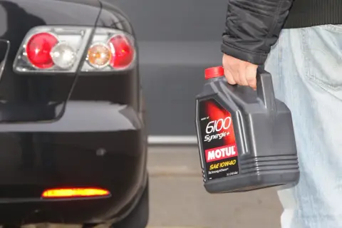Osoba trzyma olej Motul 6100 Synergie+ SAE 10W40 obok tylnej części czarnego samochodu. Idealny wybór, by dobrze dobrać olej.