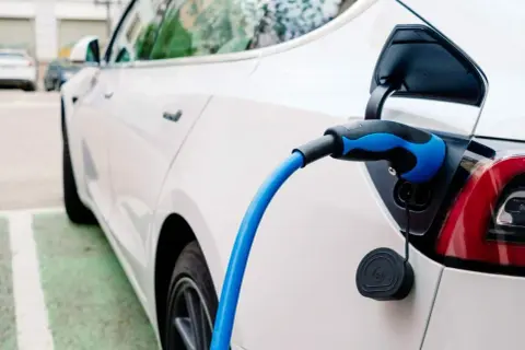 Sekrety doboru opon do aut elektrycznych: dlaczego zwykłe opony niszczą się szybciej? Biały samochód elektryczny podłączony do ładowania.