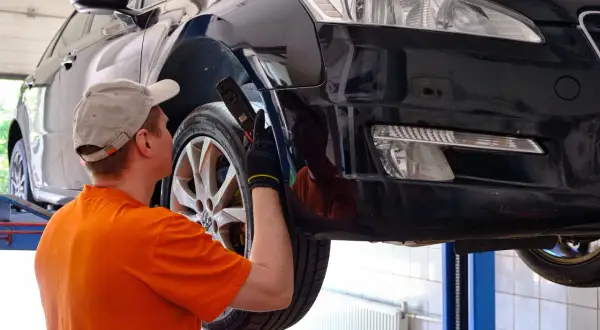 Mechanik w pomarańczowej koszulce sprawdza błotnik czarnego auta na podnośniku. Może to być powód, dlaczego mechanicy w dużych aglomeracjach rezygnują z napraw "od ręki".
