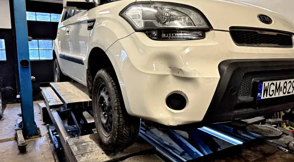 Biały Kia Soul na podnośniku, przygotowany do prac, jak wyciąć otwór na hak holowniczy w dolnej części zderzaka.