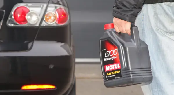 Osoba trzyma olej Motul 6100 Synergie+ SAE 10W40 obok tylnej części czarnego samochodu. Idealny wybór, by dobrze dobrać olej.