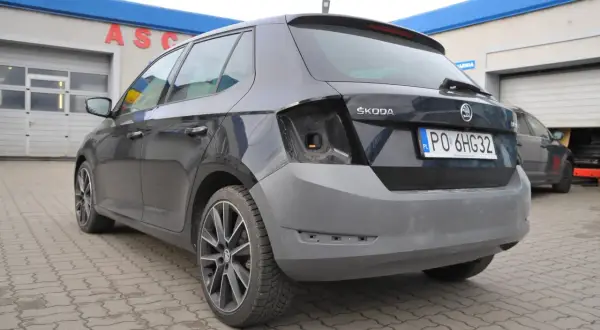 Ciemnoszara Skoda z niepomalowanym zderzakiem tył, przed warsztatem samochodowym.