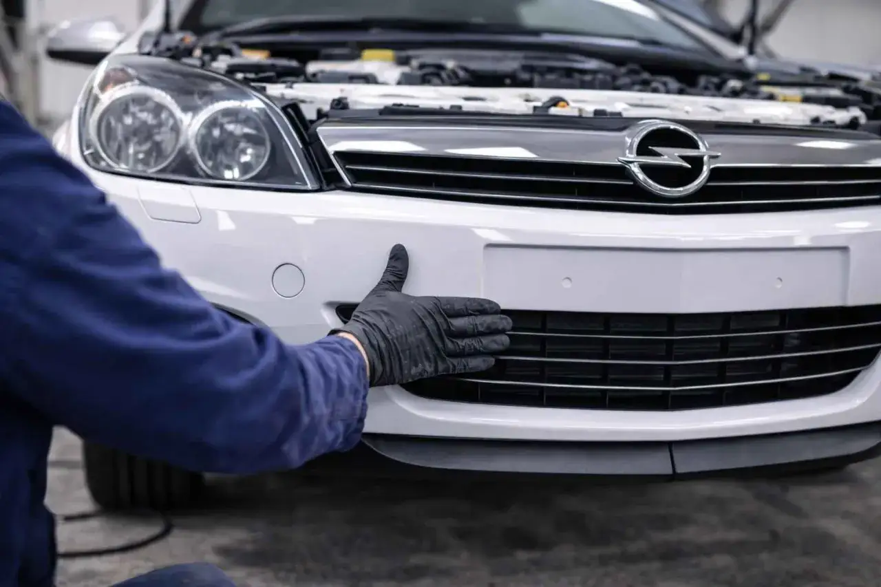 Mechanik w czarnej rękawiczce wykonuje wymianę kratek zderzaka bez zdejmowania całego elementu w białym Oplu.