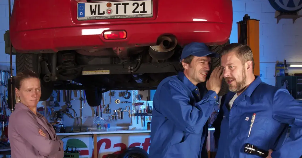 Mechanicy szepczą o prawdziwych powodach dla których serwisy odmawiają montażu części klienta, podczas gdy kobieta patrzy z dezaprobatą.