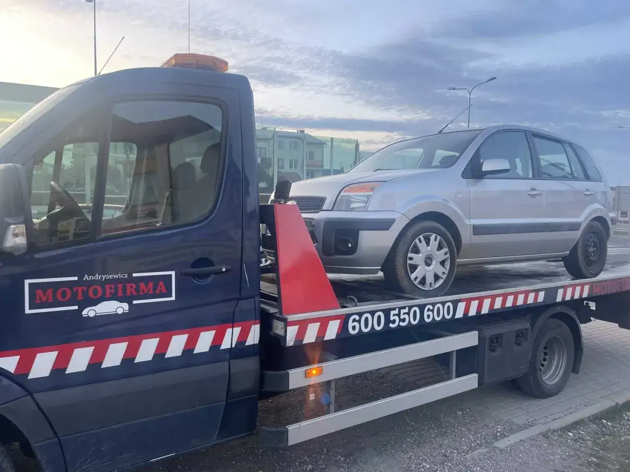 Potrzebujesz pomocy drogowej? Sprawdź, ile kosztuje laweta od firmy Motofirma. Srebrny samochód na platformie.