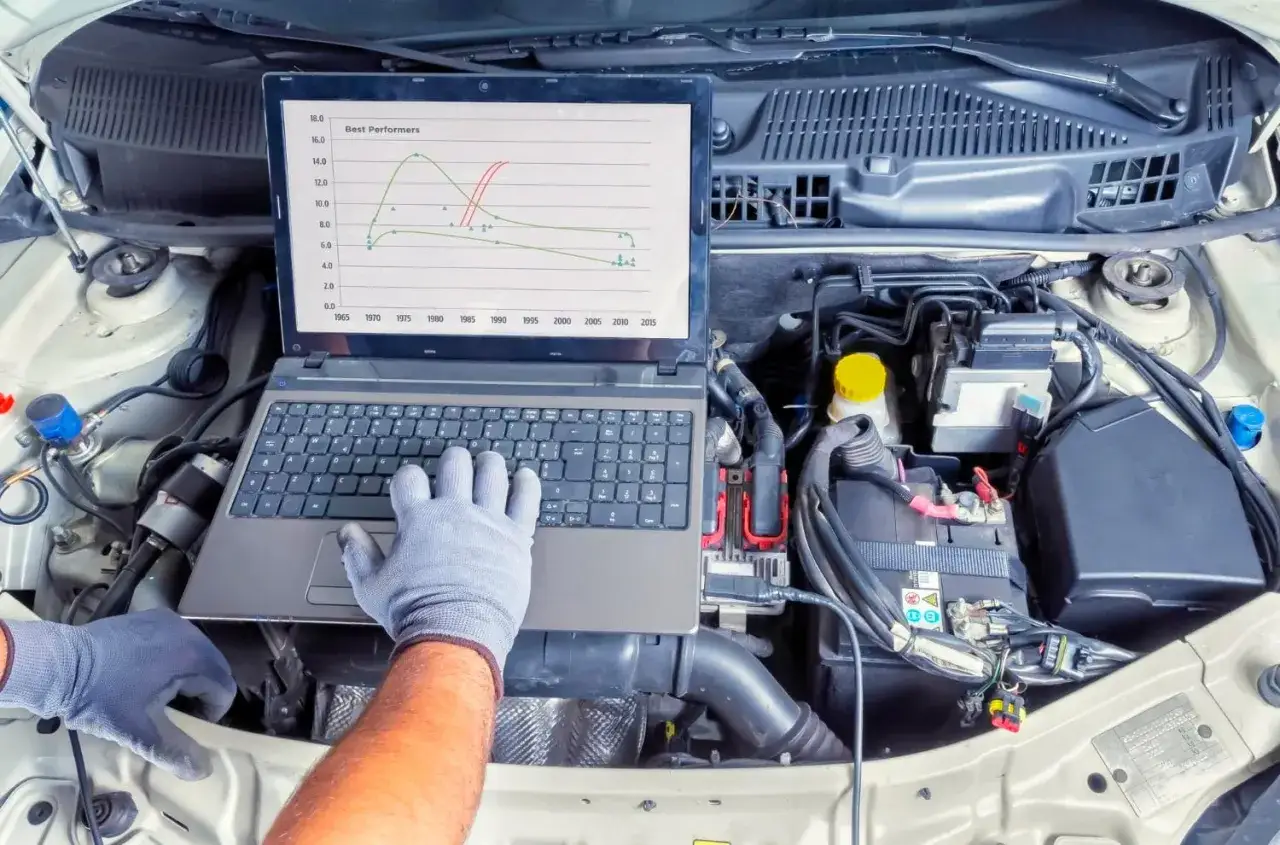 Ręce w rękawiczkach nad laptopem w komorze silnika. Wykres na ekranie może sugerować ukryte przyczyny dla których mechanik każe Ci płacić za diagnostykę komputerową dwa razy.
