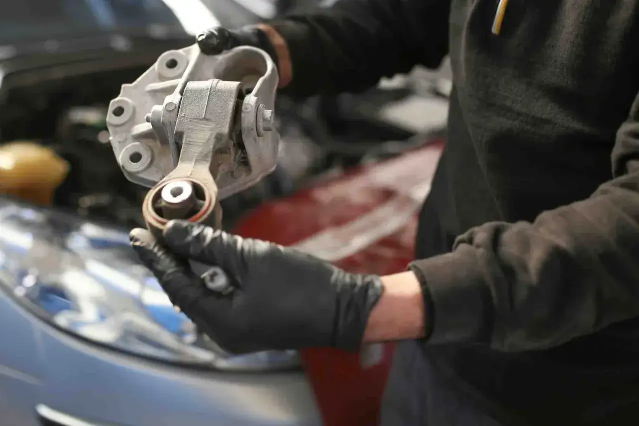 Mechanik w czarnych rękawiczkach trzyma metalowy element, który jest częścią układu amortyzującego silnik. To kluczowy element, gdy zastanawiasz się, jak sprawdzić poduszki silnika.