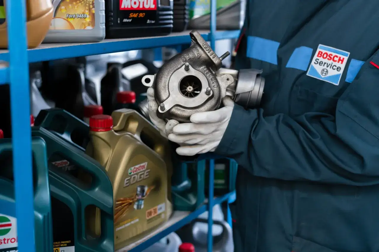 Mechanik w rękawiczkach trzyma turbosprężarkę. Na półce widać oleje silnikowe, które pomagają jak wydłużyć żywotność turbosprężarki w silniku z start-stop.
