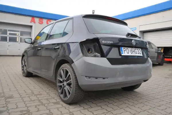 Ciemnoszara Skoda z niepomalowanym zderzakiem tył, przed warsztatem samochodowym.